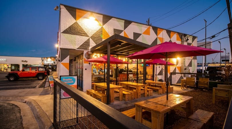 Outdoor patio of a restaurant with wooden picnic tables, red umbrellas, and a colorful geometric mural.