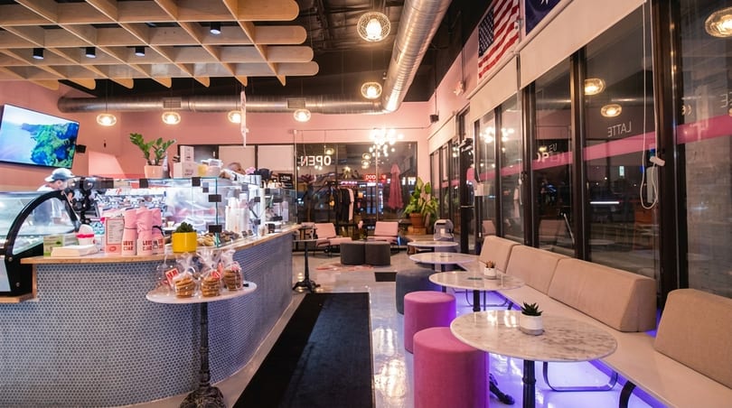 Modern cafe interior with pink walls, blue tiled counter, marble tables, and plush seating.