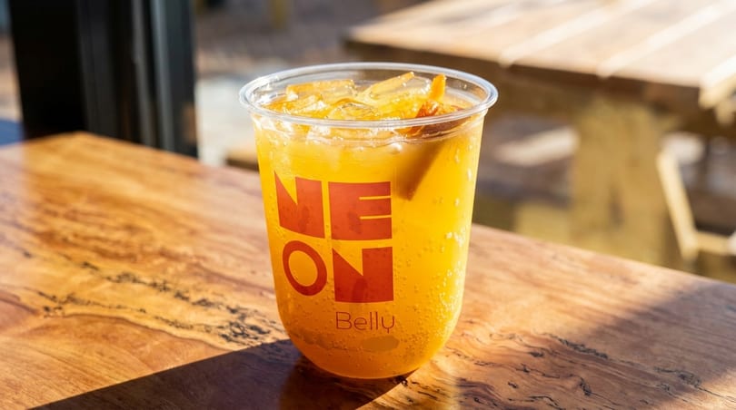 Orange drink with ice and fruit in a "NEON Belly" cup on a wooden table.