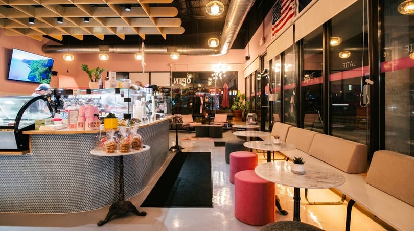 Modern cafe interior with pink walls, a long counter, and seating areas with round tables and pink stools.