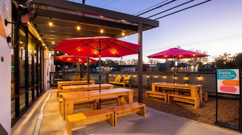 Outdoor restaurant patio with wooden picnic tables, red umbrellas, string lights, and a "Patio for Neon Belly customers" sign.