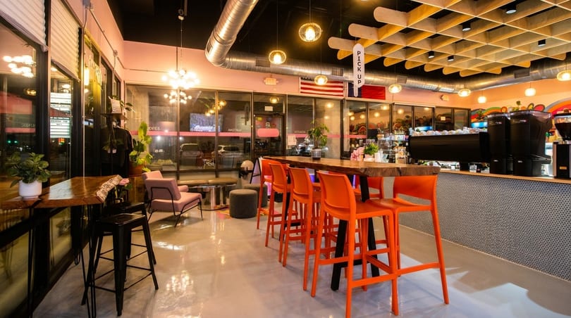 Modern cafe interior with orange bar stools, a long wooden table, and a coffee bar.