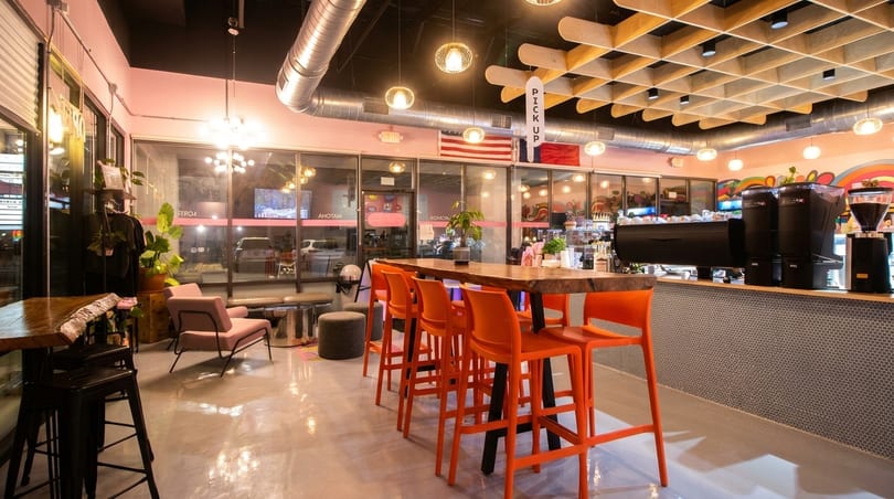 Modern cafe interior with orange bar stools, a long wooden table, and a coffee bar.